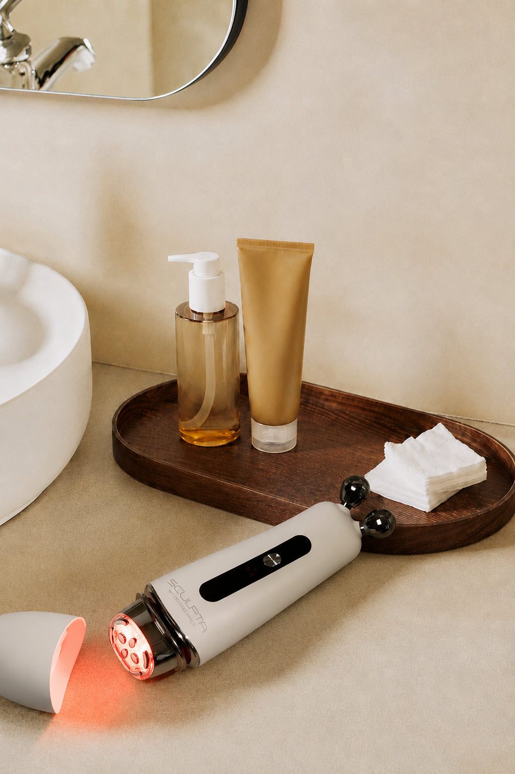 Déesse PRO Sculpta device emitting red LED light beside skincare bottles and cotton pads on wooden tray